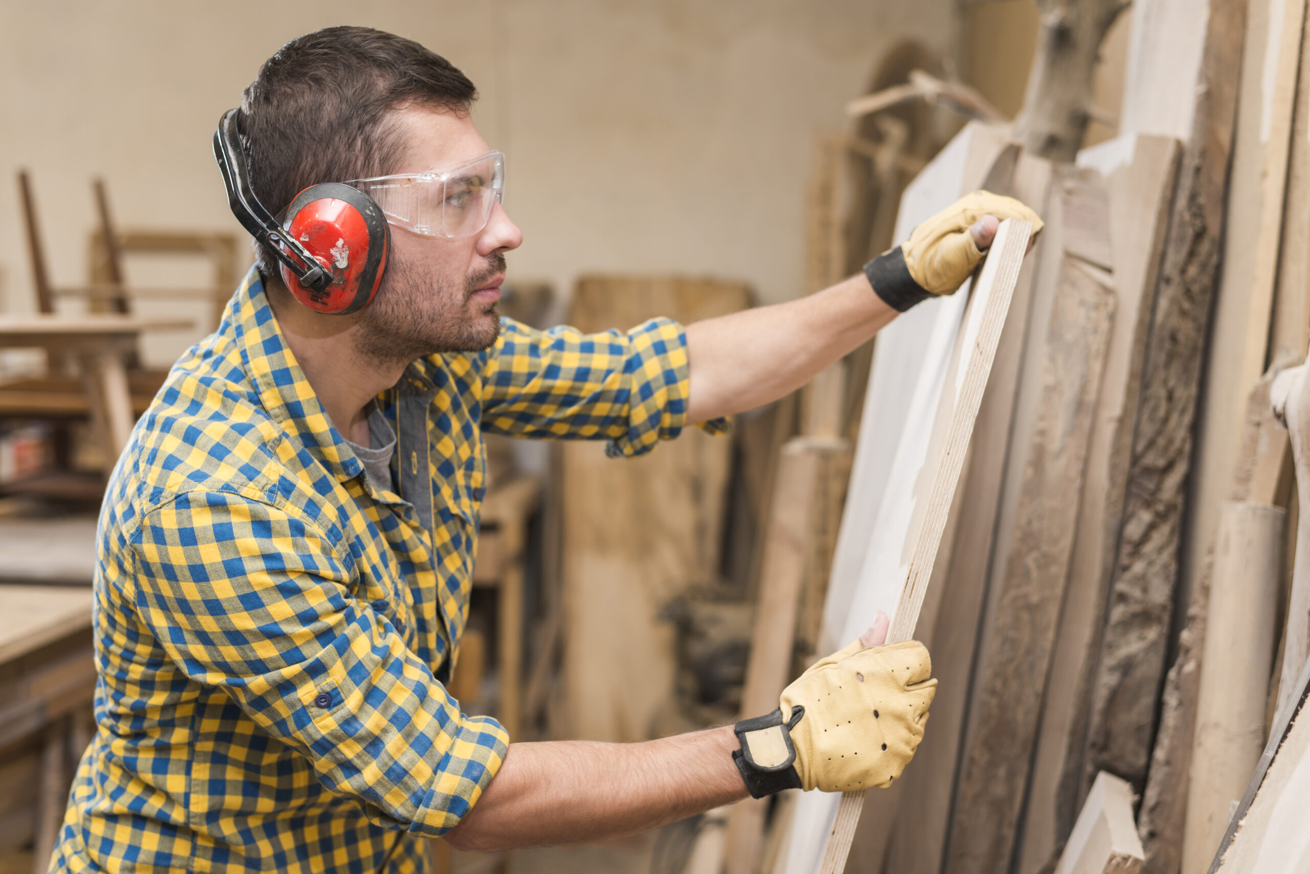 male-carpenter-wearing-safety-glasses-checking-wooden-plank-workshop